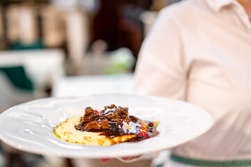 Close-up of gourmet meat dish with dark sauce, mashed base, edible flowers, and berries on white plate, held by server in blurred restaurant background