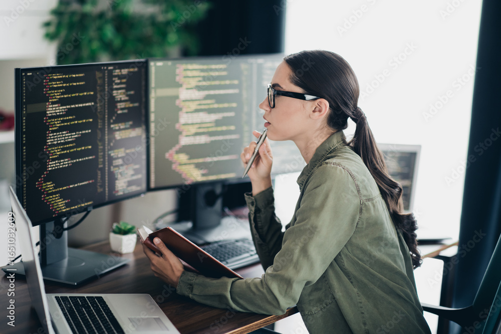 Poster focused young female programmer analyzing code on computers while working on laptop in an organized 