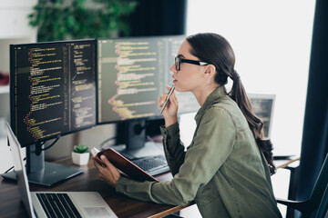 Focused young female programmer analyzing code on computers while working on laptop in an organized...