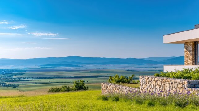 Modern house on a hilltop overlooking expansive green fields and distant mountains under a clear blue sky