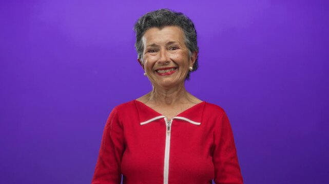 Woman smiling confidently while pointing directly at the camera, standing against a vibrant purple background, radiating positivity and energy in a colorful red outfit.