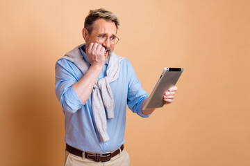 Mature businessman with stylish outfit holding a tablet and showing concern on a beige studio...