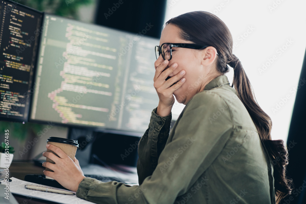Poster young female programmer yawning while working on code and holding a coffee cup in a modern workspace