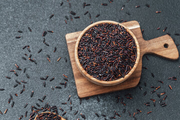 Black rice in wooden bowl with spoon and scattered grains on dark background