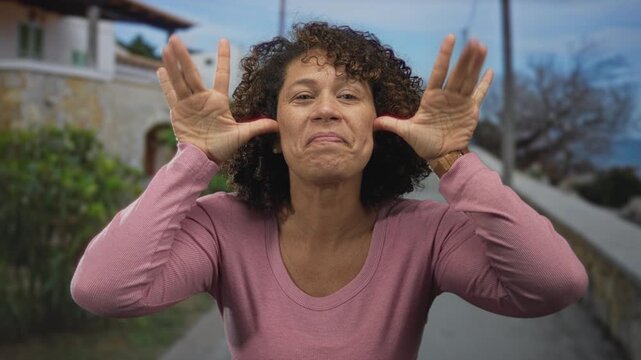 Woman in pink shirt sticks tongue out under bright sunlight with crossed arms on street; playful mischief.