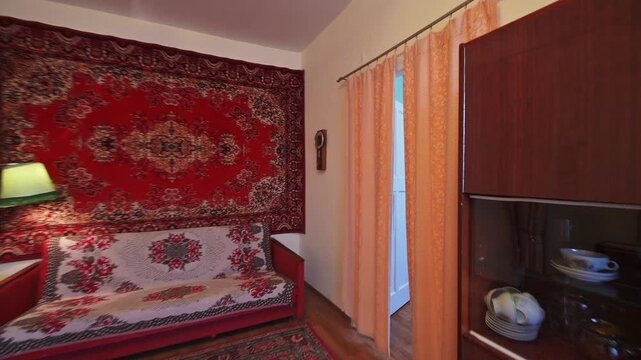 vintage living room with a rug-covered wall, wooden furniture, and a table. Light pink curtains decorate the windows.
