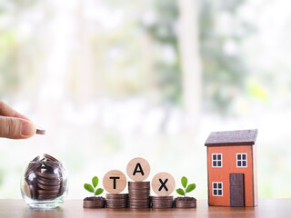 Hand putting coin into glass bottle, Miniature house, Wooden blocks with the word TAX on stack of...