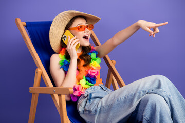 Charming young woman in summer outfit sitting on deck chair using phone on vibrant purple background