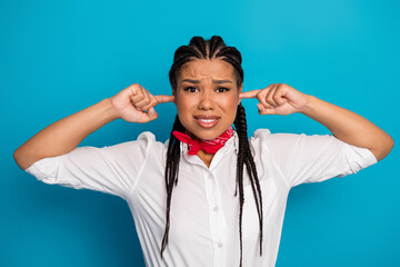 Young woman with braids plugs ears with fingers while expressing frustration on a blue background