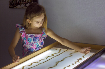 Beautiful little girl draws with sand on a light square, art therapy, sand animation, creative...