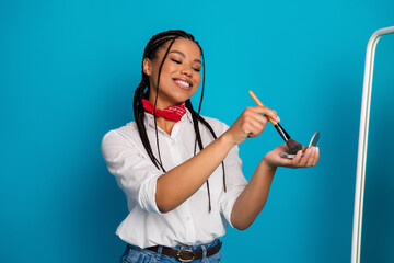 Charming young woman applying makeup with a brush in a casual white shirt against a vibrant blue...