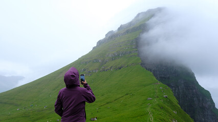 A traveler in a purple jacket stands on lush green slopes, capturing the dramatic cliffs of the...