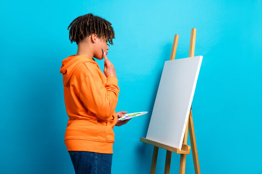 Young boy in an orange hoodie contemplating art on a blank canvas against a vibrant blue background