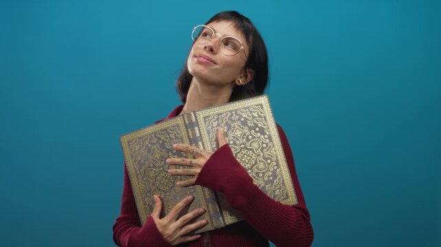 Woman reading ornate hardcover book with tattooed hand holds pages open in bright blue studio; knowledge curiosity.