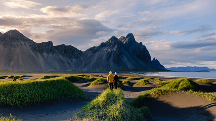 A couple strolls hand in hand across the unique black sand beach in Iceland, surrounded by lush green grasses and towering mountains. Stokksnes beach Vestrahorn mountain Iceland