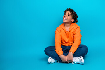 Smiling boy in an orange hoodie seated on a vibrant blue background, radiating happiness and style,...