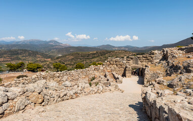 Ancient stone gateway at the Mycenae archaeological site in Greece, surrounded by rugged hills and olive groves under bright midday sunlight, blue sky