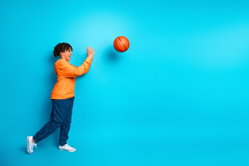 Happy schoolboy playing with a basketball against a bright blue background wearing trendy casual orange hoodie and jeans