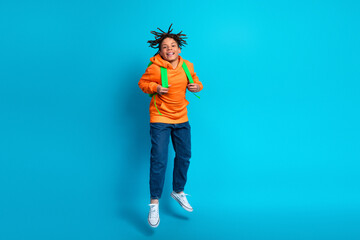 Cheerful young boy jumping with a backpack against a blue background wearing an orange hoodie and jeans