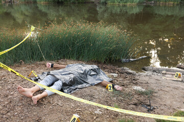 Forensic expertise. Crime scene with dead man's body, markers and police tape near lake outdoors