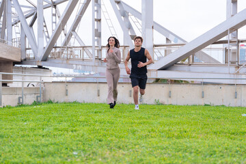 Young man and woman jogging together outdoors on green grass under a large metal bridge, wearing...