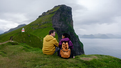 A couple sits closely on a grassy ledge, gazing at the breathtaking cliffs of the Faroe Islands....