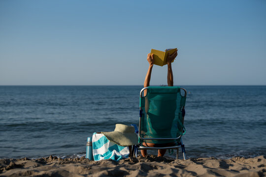 Rear view of woman sitting on beach chair reading yellow book with arms raised, relaxing by calm sea with straw hat, towel, and thermos beside her on sunny summer morning - Powered by Adobe