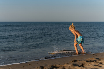 Shirtless young blond boy with hair in a bun playing at the shoreline, wearing blue floral shorts,...