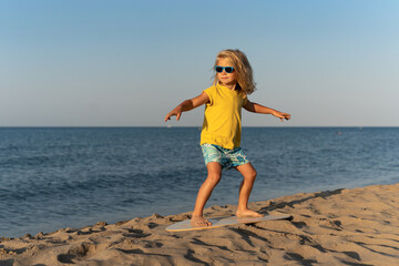Blond boy wearing yellow t-shirt and blue patterned shorts balancing barefoot on a skimboard on...