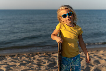 Smiling blond child in yellow t-shirt and blue patterned shorts standing barefoot on sandy beach,...