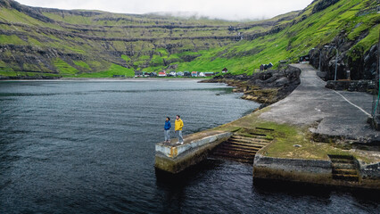 Two travelers stand on a tranquil pier in the Faroe Islands, surrounded by lush green hills and quaint village buildings. Tjornuvik village