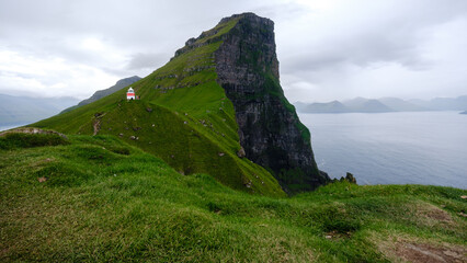 Emerging from lush green hills, a solitary lighthouse stands proudly atop dramatic cliffs overlooking the tranquil sea in the Faroe Islands. Kallur Lighthouse, Kalsoy Island