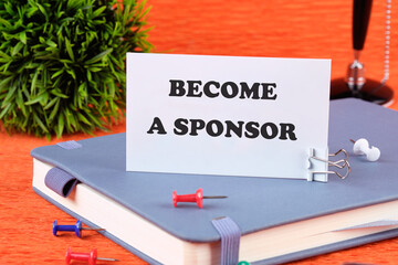 BECOME A SPONSOR text written on a business card standing with a clip on a diary, a notebook on an...