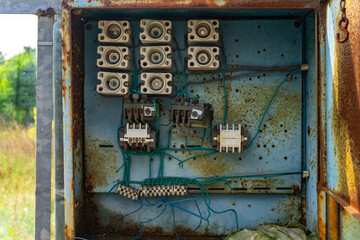 Rusty electrical box with porcelain fuses and tangled wires, mounted on a blue metal background, located in an abandoned industrial area.
