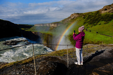 A visitor stands in awe, capturing the breathtaking Gullfoss waterfall as a vibrant rainbow appears...