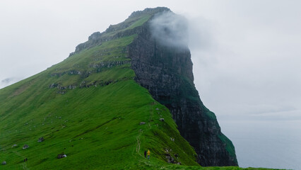 A lone hiker traverses the verdant slopes of the Faroe Islands, surrounded by towering cliffs and shrouded in mist, capturing the wild beauty and rugged terrain, Kallur Lighthouse Kalsoy Island