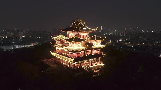 Hangzhou, China. A nighttime aerial view of a traditional Chinese pagoda, illuminated in gold and pink hues, set against a backdrop of a cityscape.