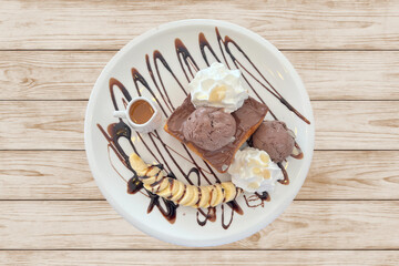 Delicious dessert plate with chocolate toast topped with scoops of chocolate ice cream, whipped cream, sliced banana drizzled with chocolate syrup, and a small jug of honey on white plate