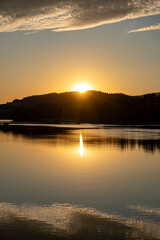 Obraz premium Bright sun setting behind forested hills, golden rays reflecting on calm lake water with evening clouds above horizon.