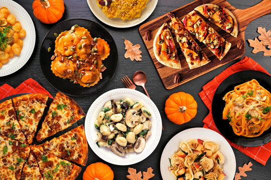 Delicious autumn meal table scene. Overhead view on a black wood background. Pumpkin and apple theme risotto, pasta, gnocchi, flat bread and pizza.