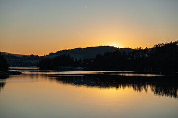 Golden sunset behind hills reflecting on calm lake water with dark forest silhouettes and peaceful sky.
