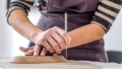 Obraz premium A close-up shot of a middle-aged woman artist shaping clay with sculpting tools at a table in an art studio, with an environment adorned with artistic paintings in the background