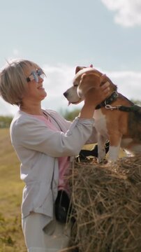 Elderly woman in sunglasses lovingly rubs her beagle ears while it sits on a haystack in a vast countryside field, another person in sunglasses is nearby