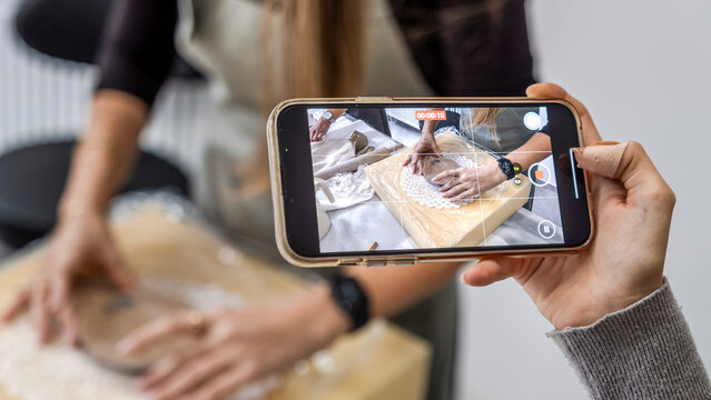 In an art studio a beautiful middle-aged woman artist is shaping clay at a table, while a young woman next to her is taking a photo with her phone - Powered by Adobe
