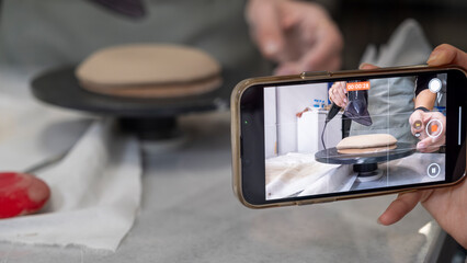 In an art studio a beautiful middle-aged woman artist is shaping clay at a table, while a young woman next to her is taking a photo with her phone