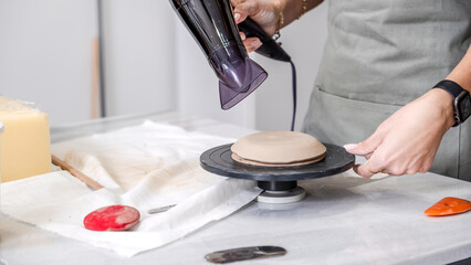 In an art studio with an environment adorned with artistic paintings in the background, a beautiful middle-aged woman artist is drying a clay-shaped plate with a hairdryer at a table