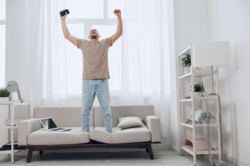 Energetic man celebrating success while standing on a sofa in a bright, modern living room. His...