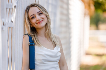 Young woman in casual attire smiles while leaning against a white fence in a sunny outdoor setting