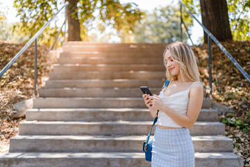 Young woman smiles while texting on her phone by the steps in a sunlit park during daytime
