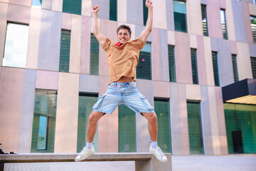 Joyful young man wearing casual clothes and headphones jumps high with arms up in the air in front...
